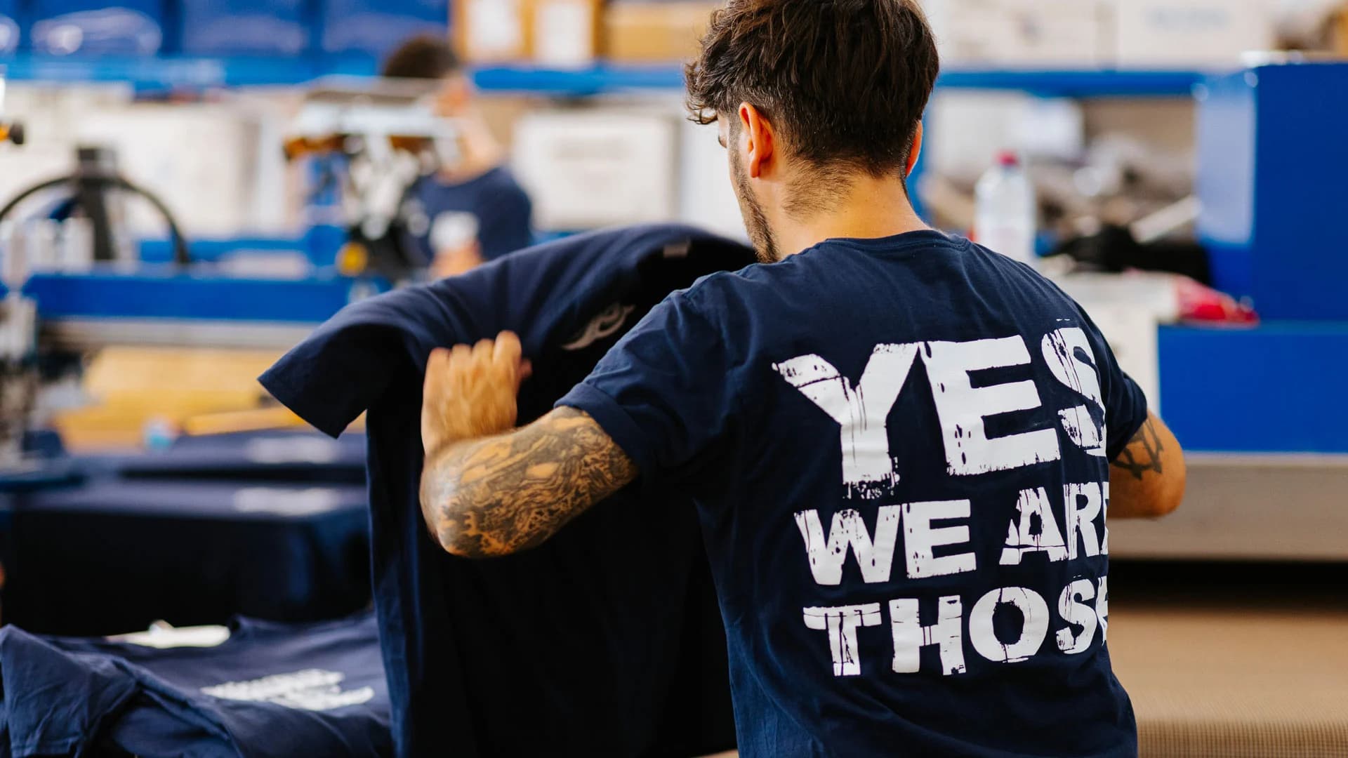 Real Pimp team member in workshop with 'Yes We Are Those We Are The Real Pimp' text on back of navy t-shirt